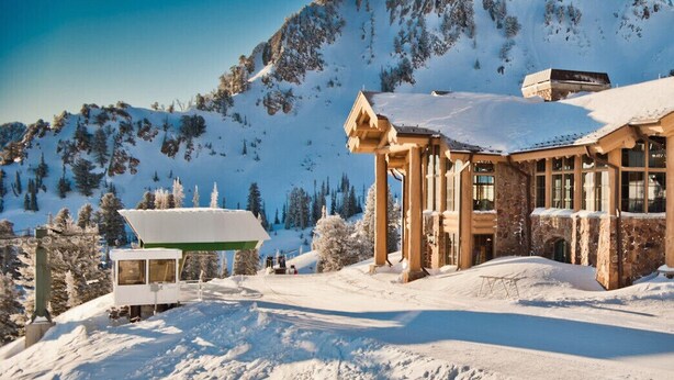 Un albergue cubierto de nieve con montañas al fondo, un telesilla y un cielo despejado.
