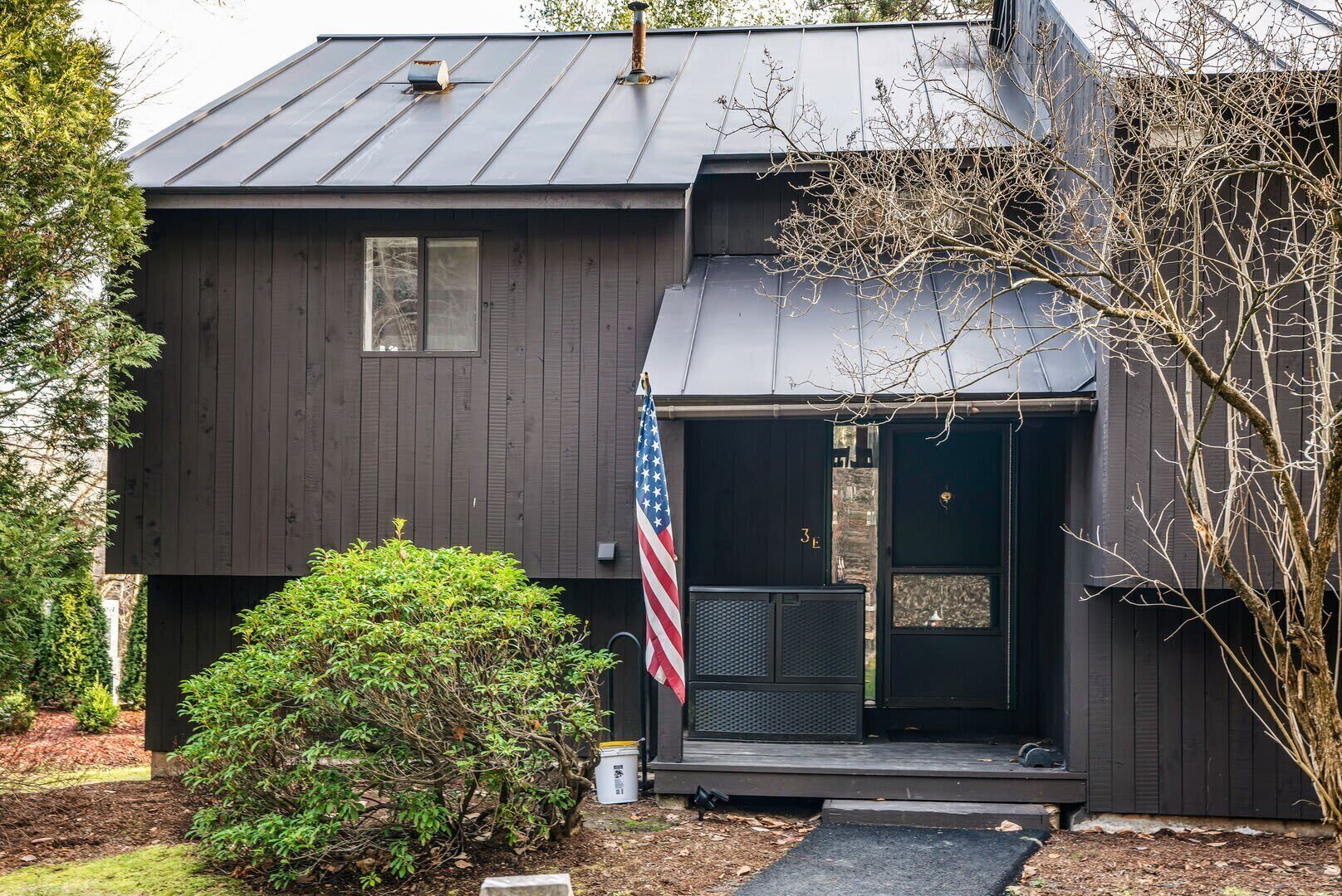 A dark wooden house with a metal roof, a small porch, and an American flag.