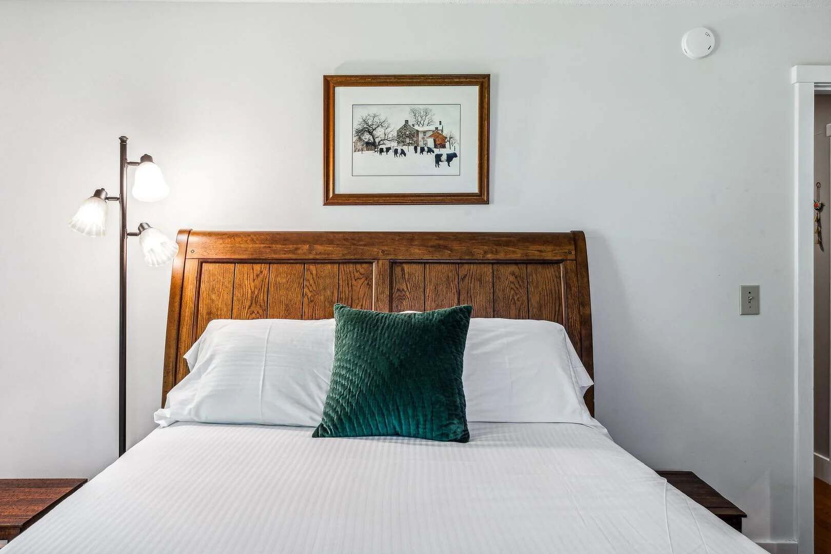 A neatly made bed with white linens and a green pillow, a wooden headboard, a framed picture on the wall, and a floor lamp.