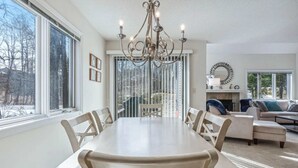 A dining area with a large table and chairs, a chandelier, and a view of the outdoors through large windows. - Condo with BBQ, Newton Village (Quechee)