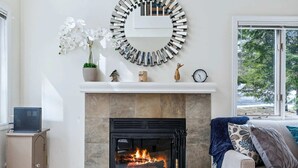 A living room with a fireplace, a white mantel, and a view of the outdoors through a large window. - Condo with BBQ, Newton Village (Quechee)
