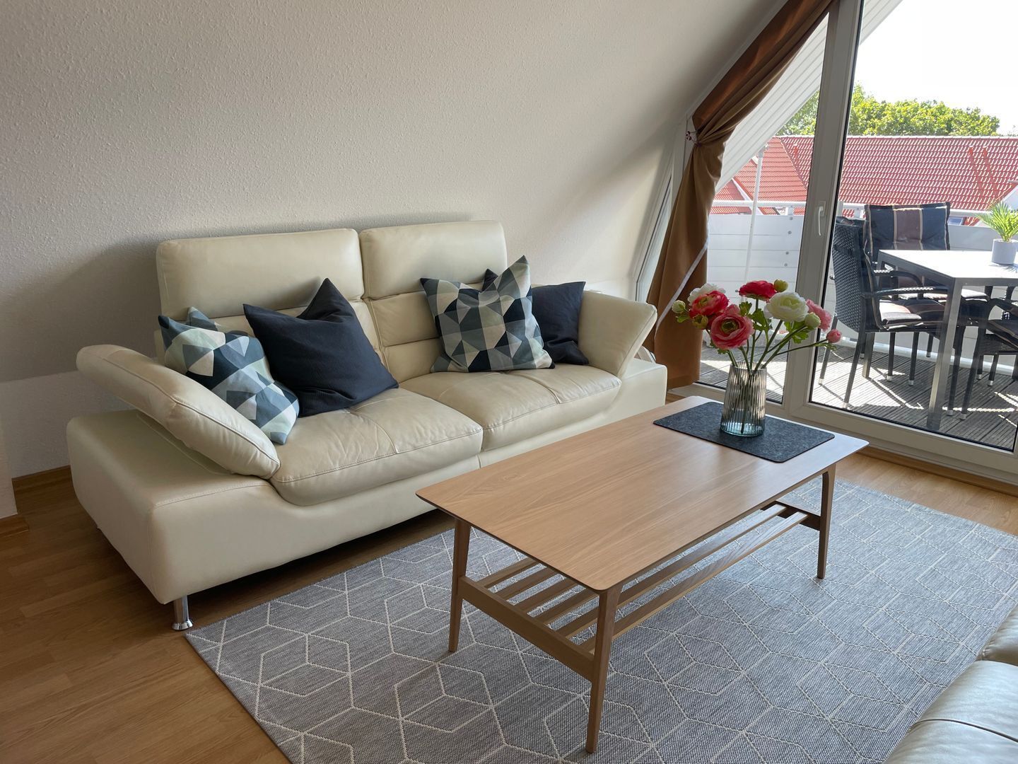 Ein Wohnzimmer mit einem beigefarbenen Sofa, gemusterten Kissen, einem Holztisch und Blick ins Freie durch ein großes Fenster.