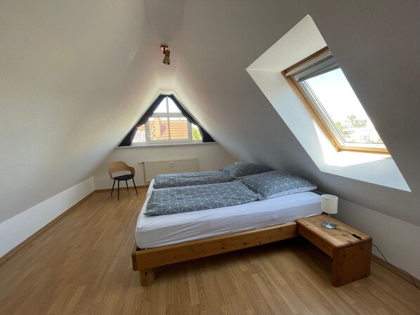 Une chambre avec un plafond en pente, un lit avec un cadre en bois, une chaise, une table de chevet avec une lampe et une fenêtre donnant sur l’extérieur.