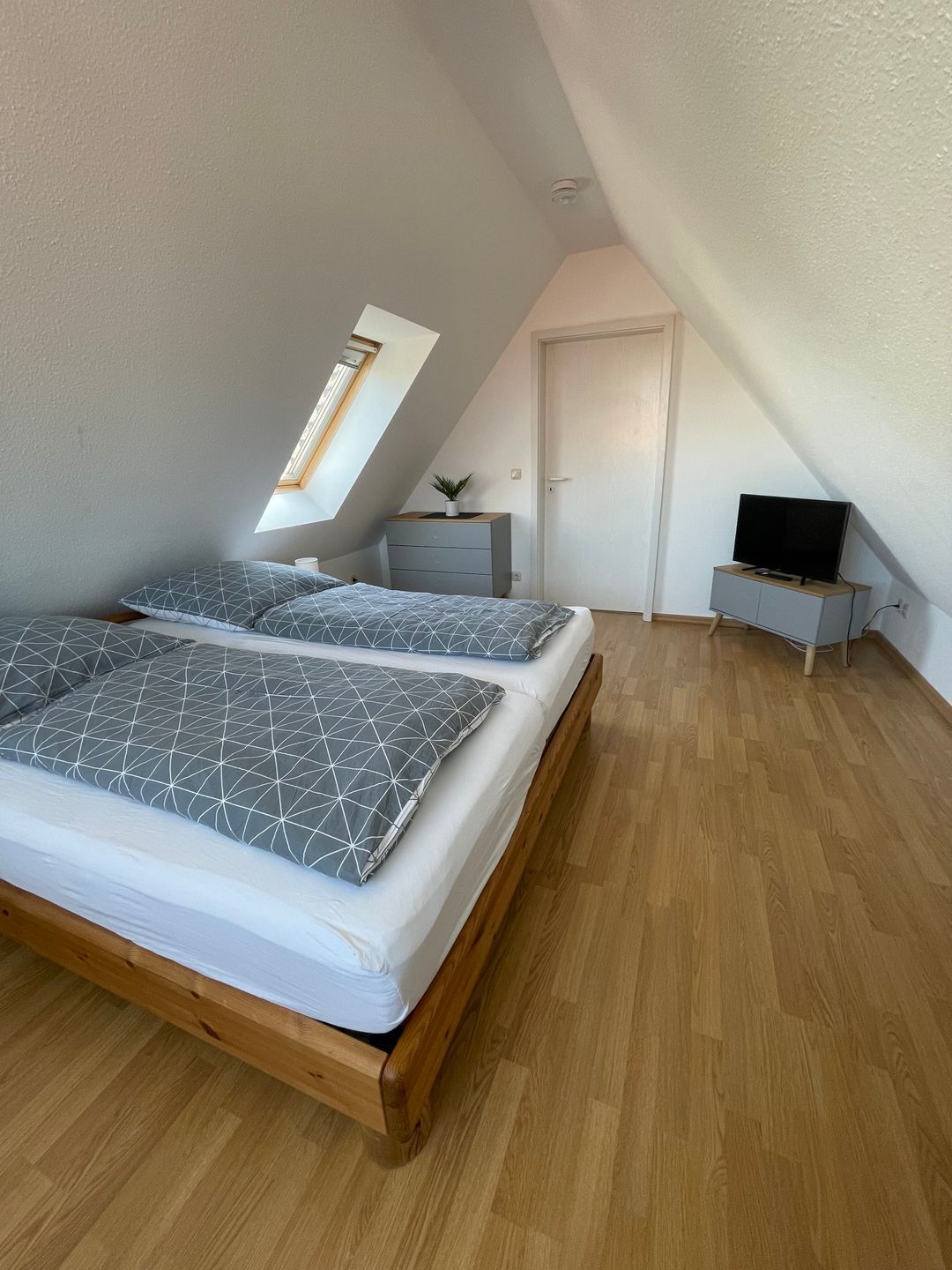 Un dormitorio con techo inclinado, una cama con almohadas de estampado geométrico, una mesita de noche pequeña, un televisor y una puerta.