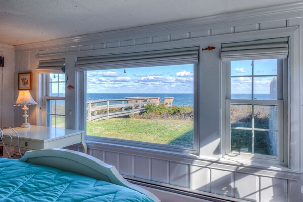 A bedroom with a large window overlooking a wooden deck and the ocean.