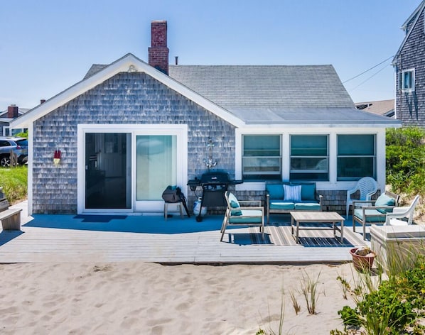 Ein Strandhaus mit einer Terrasse, Sitzgelegenheiten im Freien und einem großen Schiebeglastür.