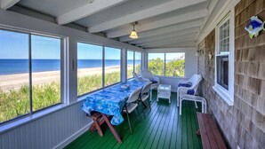 Een overdekt buitengebied met een tafel en stoelen, grote ramen met uitzicht op het strand en een houten terras.
