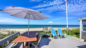 Een houten terras met een tafel en stoelen, met uitzicht op een strand en een heldere hemel, met een Amerikaanse vlag.