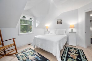 A bedroom with a bed, two bedside lamps, a rocking chair, a window, and a patterned rug. - Sun & Serenity at A Wave From It All (Pocasset)
