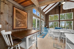Une pièce spacieuse avec un plafond en bois, de grandes fenêtres, un coin repas avec des chaises blanches et un tableau accroché au mur.