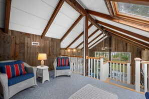 A cozy room with a sloped ceiling, wooden beams, a white railing, and a view of the outdoors. - Sun & Serenity at A Wave From It All (Pocasset)