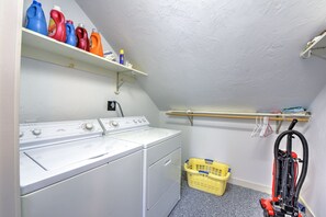 A laundry room with a washing machine, dryer, shelves with detergent bottles, a yellow laundry basket, and a vacuum cleaner. - Sun & Serenity at A Wave From It All (Pocasset)