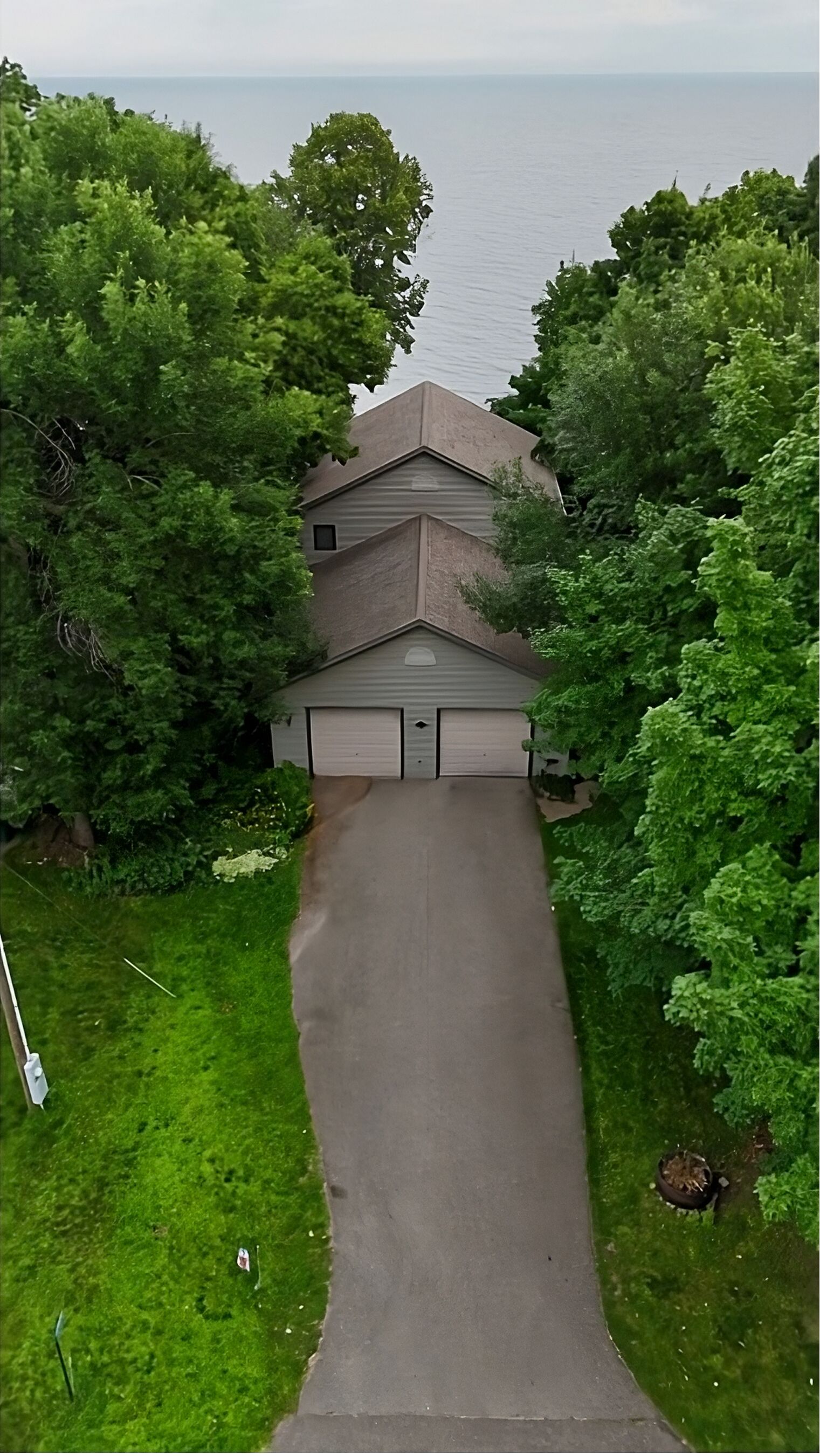 An aerial view of a house with a garage, surrounded by lush green trees and a clear view of the water body in the background.