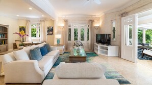 Amplia sala de estar con sofá blanco, una mesa de centro y televisor. Se aprecia una estantería y un jarrón decorativo con flores.