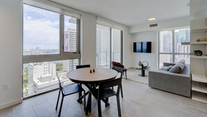 A modern living room with a round table and chairs, a grey sofa, and a TV mounted on the wall. Large windows offer a city view.