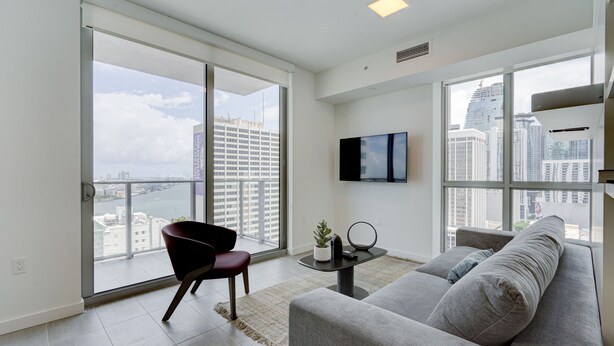 A modern living room with a large sliding glass door leading to a balcony, a grey sofa, a black chair, a small table with a plant, and a flat-screen TV mounted on the wall.