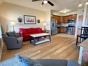 Ein Wohnzimmer mit einem roten Sofa, einem gestreiften Sessel und einem Couchtisch. Im Hintergrund ist eine Küche mit Holzschränken und Edelstahlgeräten zu sehen.
