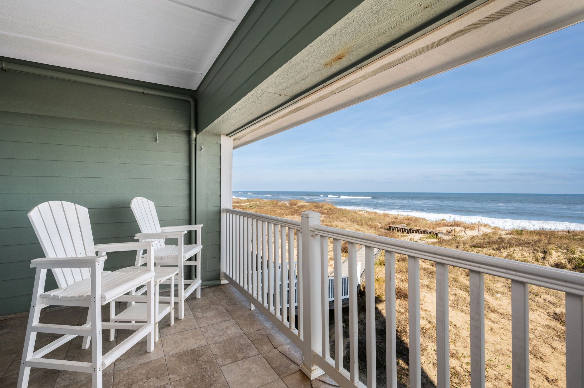 A balcony with white Adirondack chairs and a view of the beach.