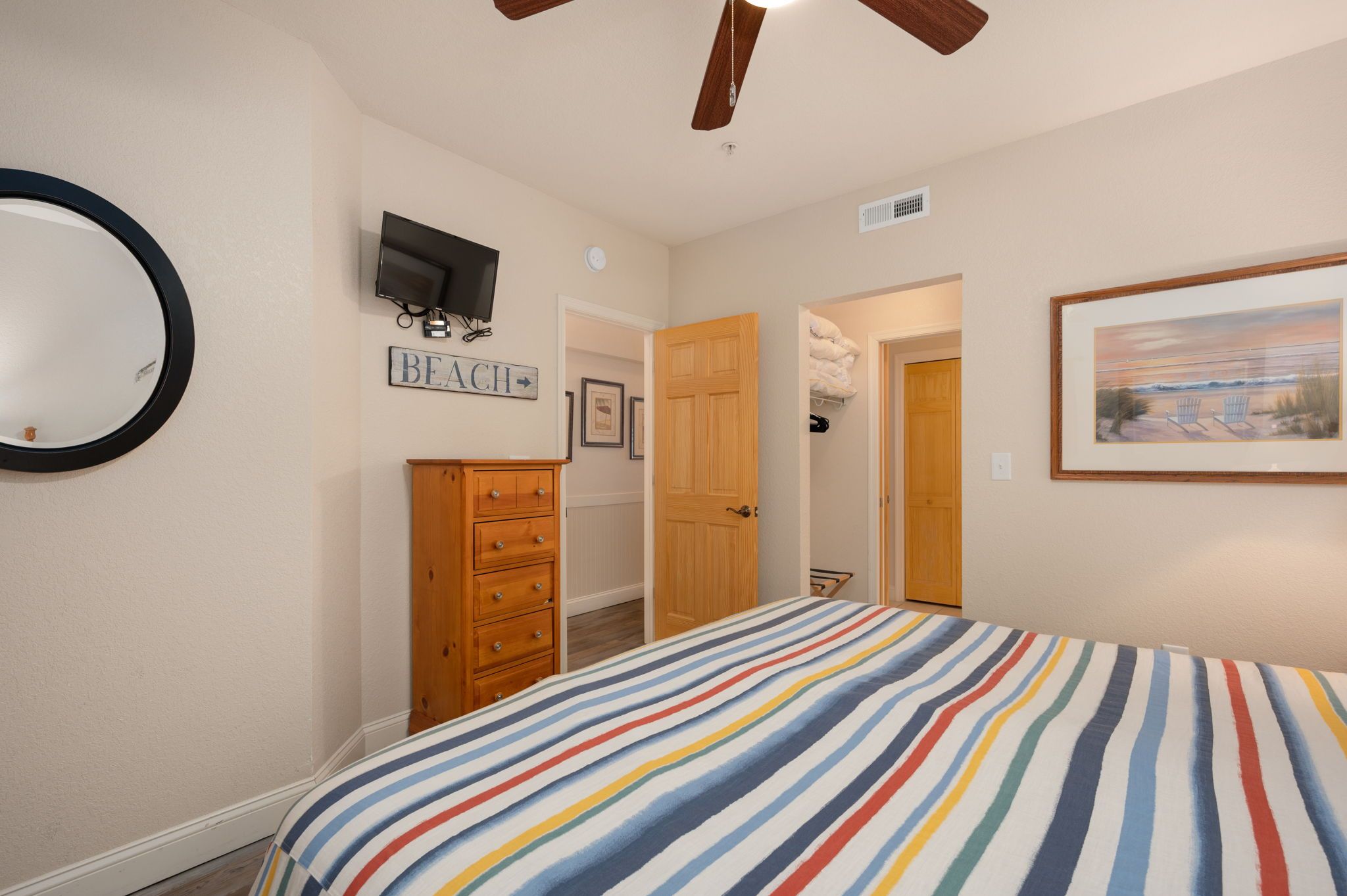 A bedroom with a striped bedspread, a wooden dresser, a television on the wall, and a door leading to another room.
