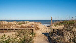 Un sentier de sable qui longe la plage et mène à l’océan, bordé de structures en bois et d’une végétation clairsemée.