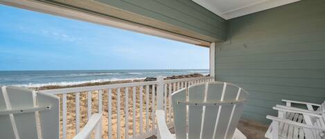 A covered outdoor area with white Adirondack chairs facing a beach.