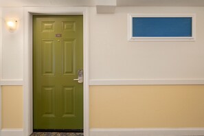 Una puerta verde con cerradura electrónica, un pequeño ventanal arriba y una lámpara en la pared a la izquierda.