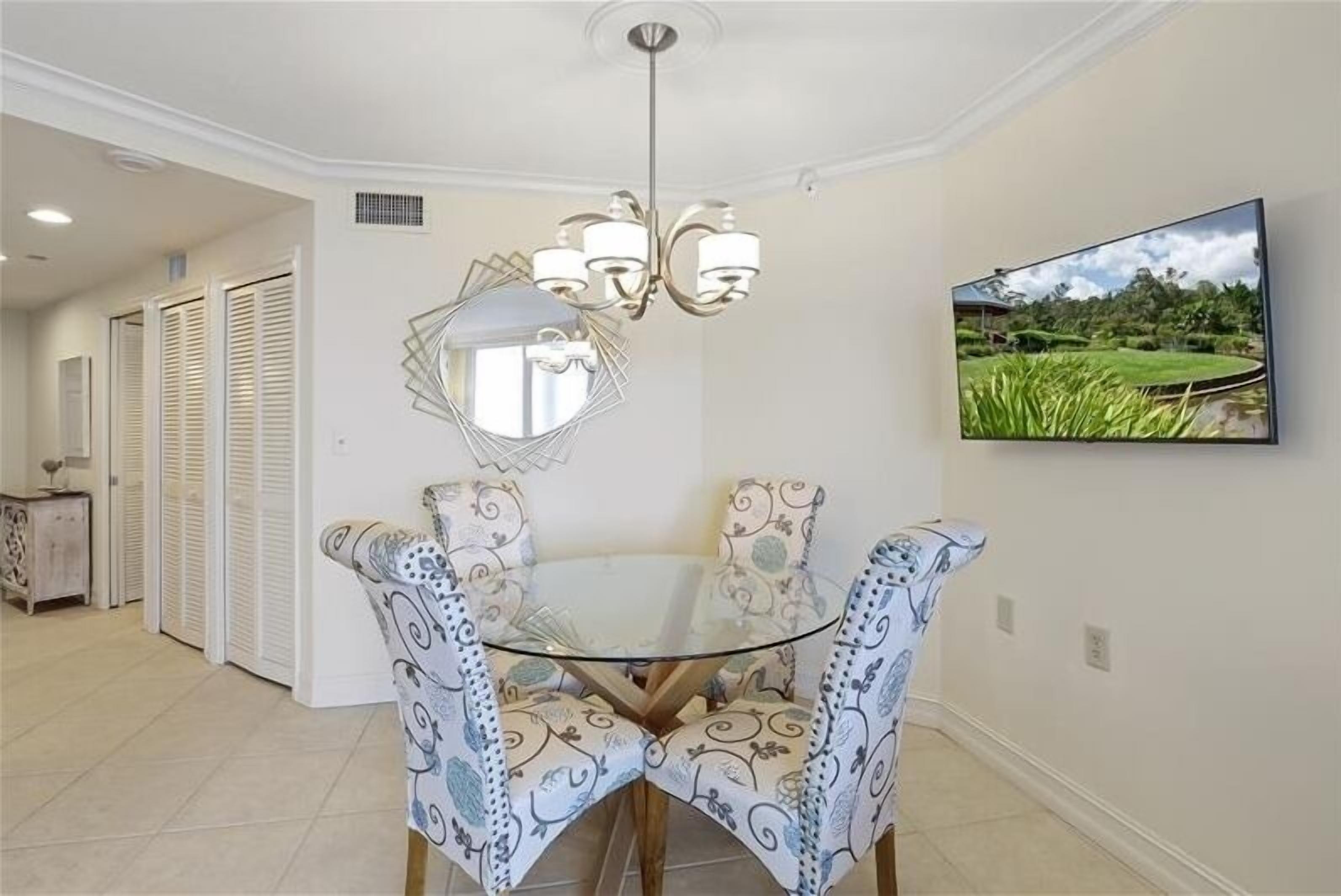 A dining area with a glass table and patterned chairs, a chandelier, and a TV displaying a landscape.