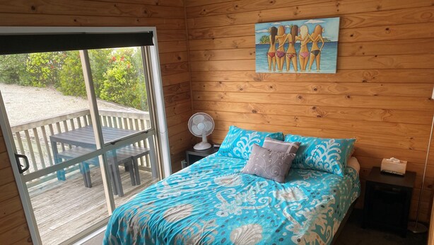 Quarto com cama, ventoinha, mesa de cabeceira e pintura de uma paisagem praiana.
