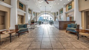 Een ruime lobby met een hoge plafond, grote ramen en een centraal gelegen zitgedeelte met banken en tafels.