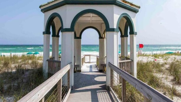 Una pérgola en la playa con techo de tejas y una terraza de madera que la conecta.