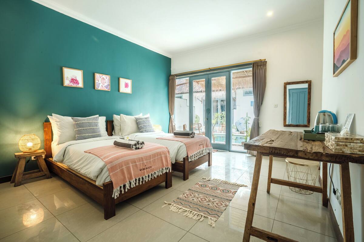 A bedroom with two beds, a wooden table, a nightstand, and a large window with curtains.
