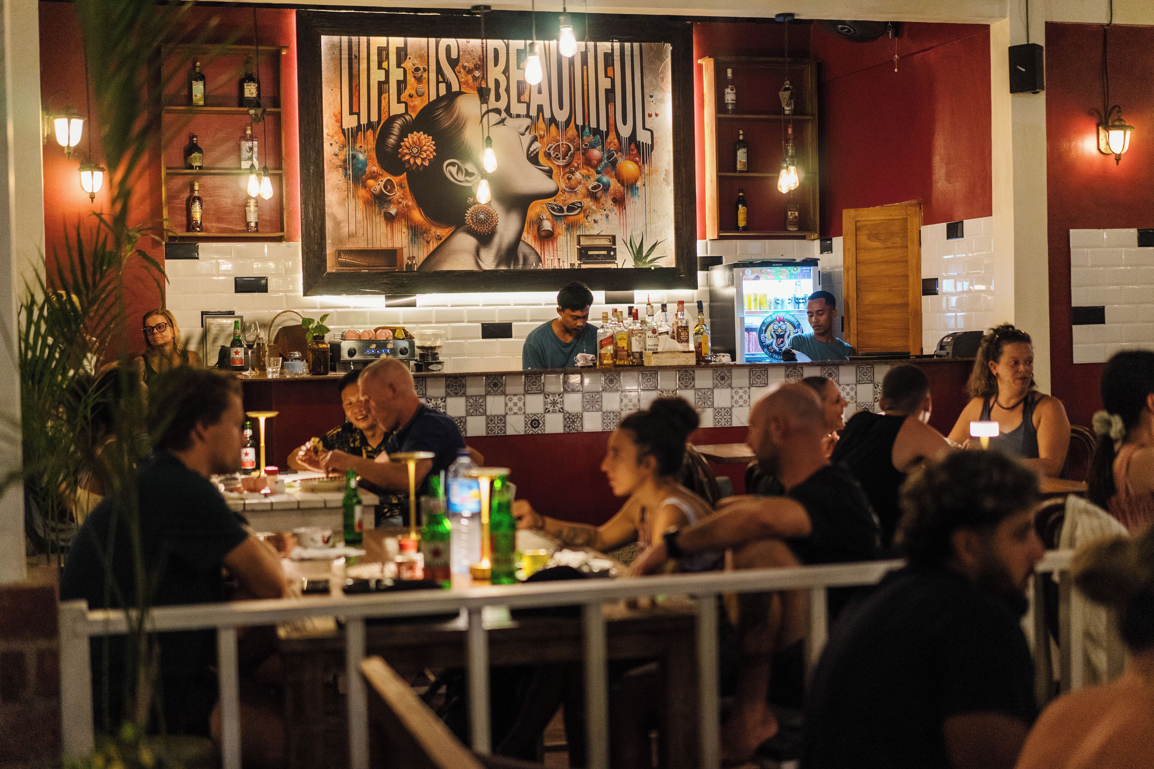 Interior de restaurant amb zona de bar, clients dinant i un mural gran a la paret.