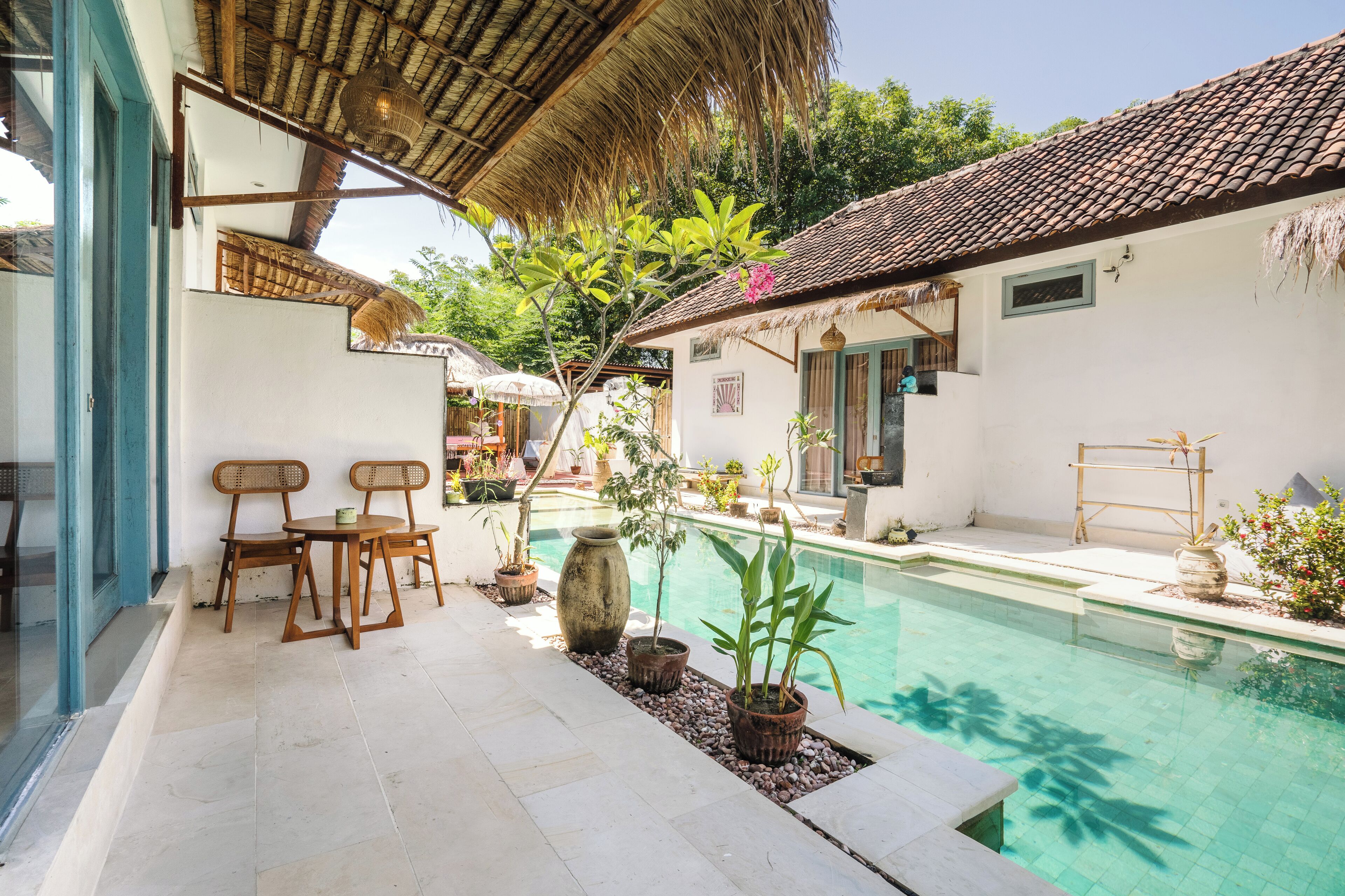 A pool area with a tiled floor, wooden chairs, potted plants, and a thatched roof structure.