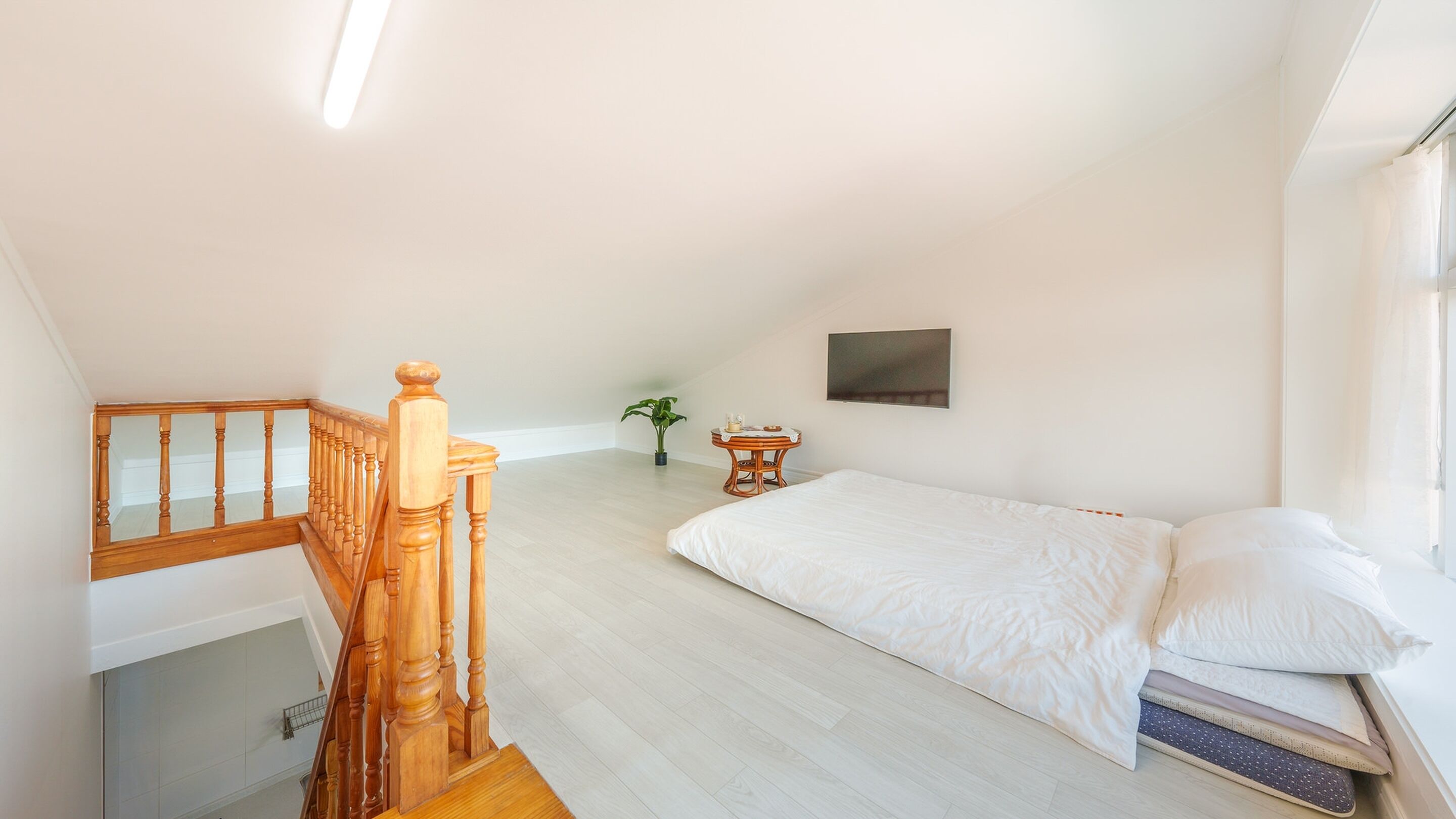 Un dormitorio con escalera de madera, una cama con ropa blanca, una mesita con una planta y un televisor montado en la pared.