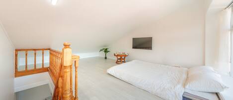 Un dormitorio con escalera de madera, una cama con ropa blanca, una mesita con una planta y un televisor montado en la pared.