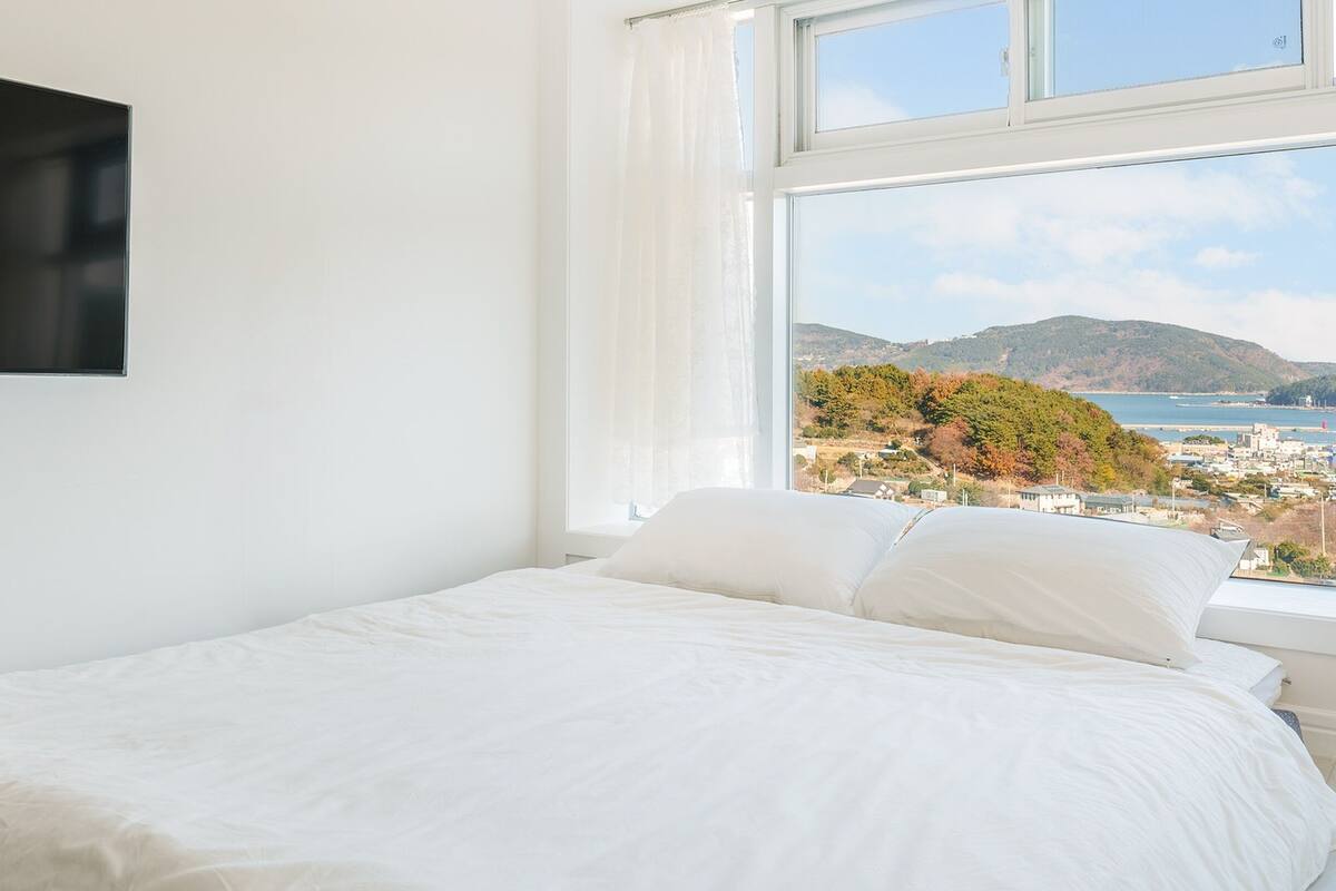 A bedroom with a bed, a TV mounted on the wall, and a window with a view of a coastal town and hills.