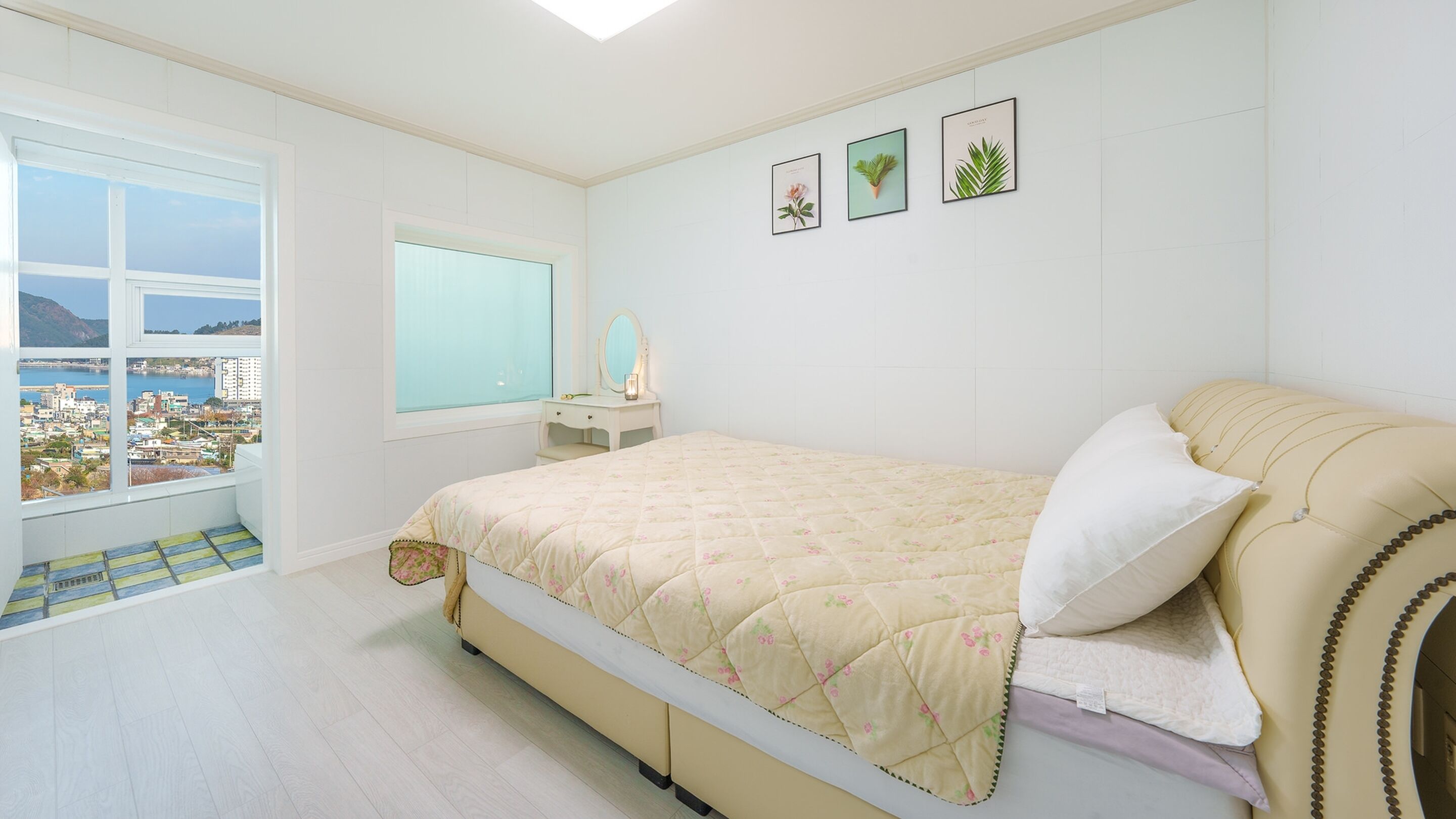 Ein Schlafzimmer mit Bett, einem Fenster mit Stadtblick, einem Nachttisch mit Spiegel sowie drei gerahmten Pflanzenmotiven an der Wand.