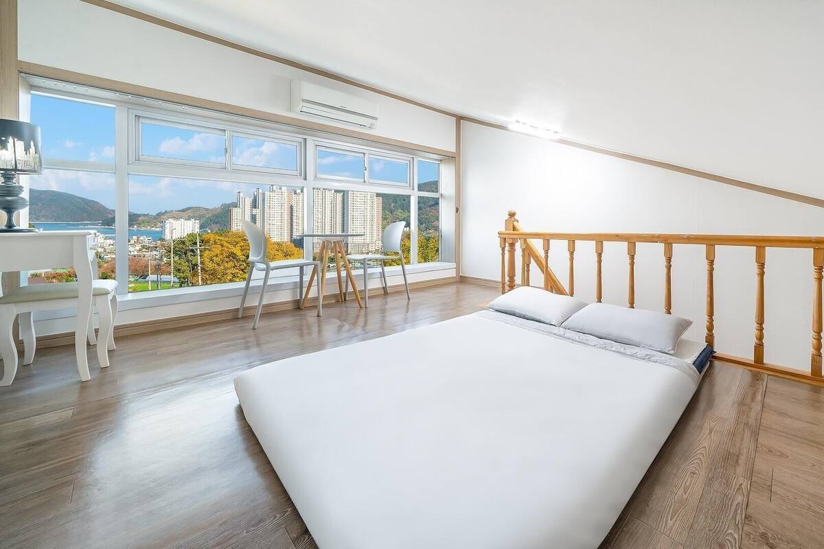 A bedroom with a large bed, a desk with a chair, and a view of a cityscape through large windows.