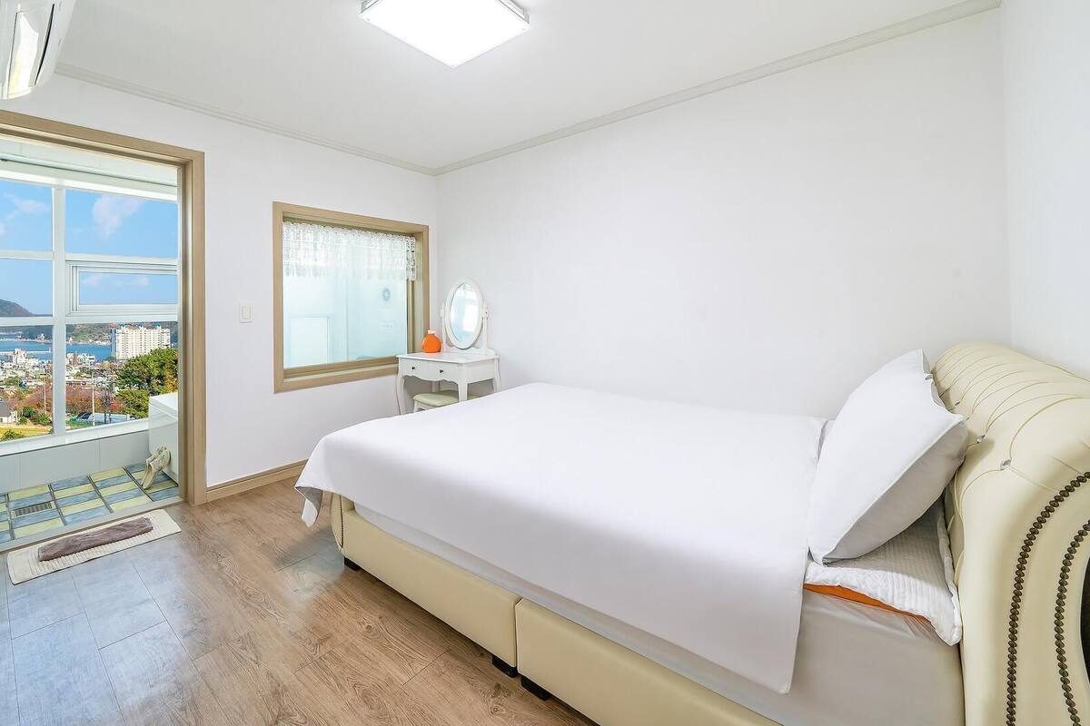 A bedroom with a bed, a bedside table, a window, and a view of the city.