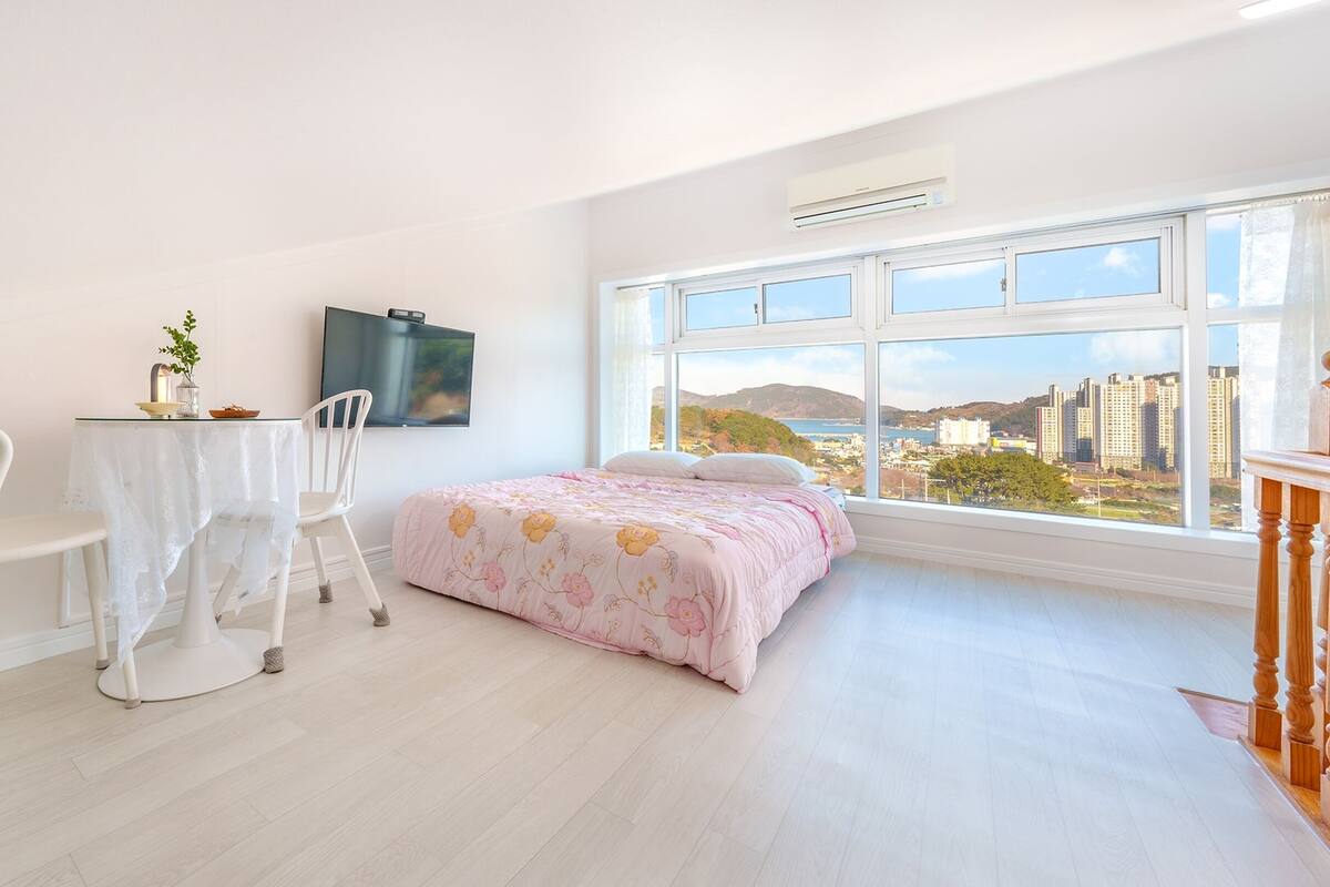 A bedroom with a bed, a desk with a chair, a TV, and a large window with a city view.