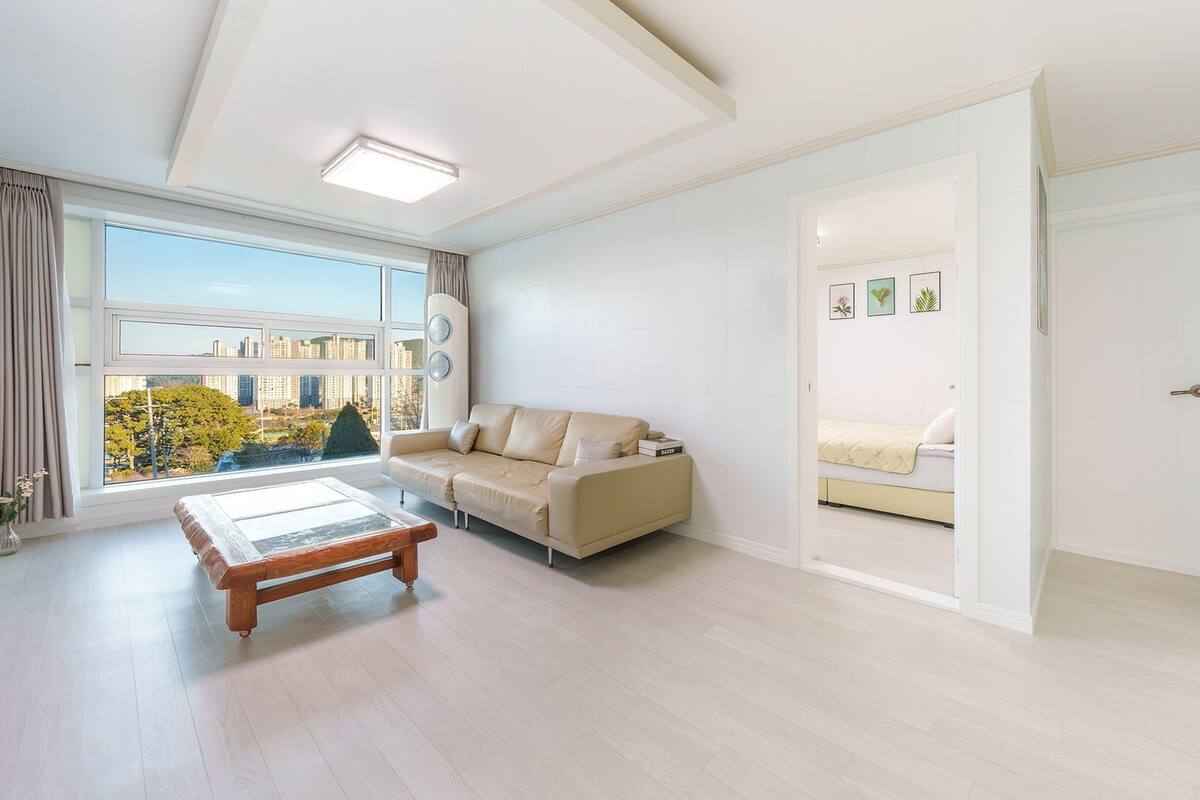 A spacious living room with a large window, a sofa, a coffee table, and a bed in the adjacent room.
