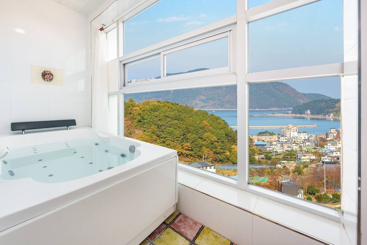 A modern bathroom with a large bathtub and a view of a coastal town through a large window.