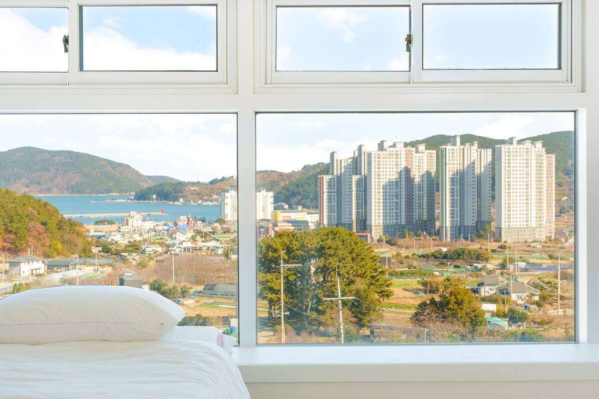 A bedroom with a large window overlooking a coastal cityscape with high-rise buildings and a body of water.