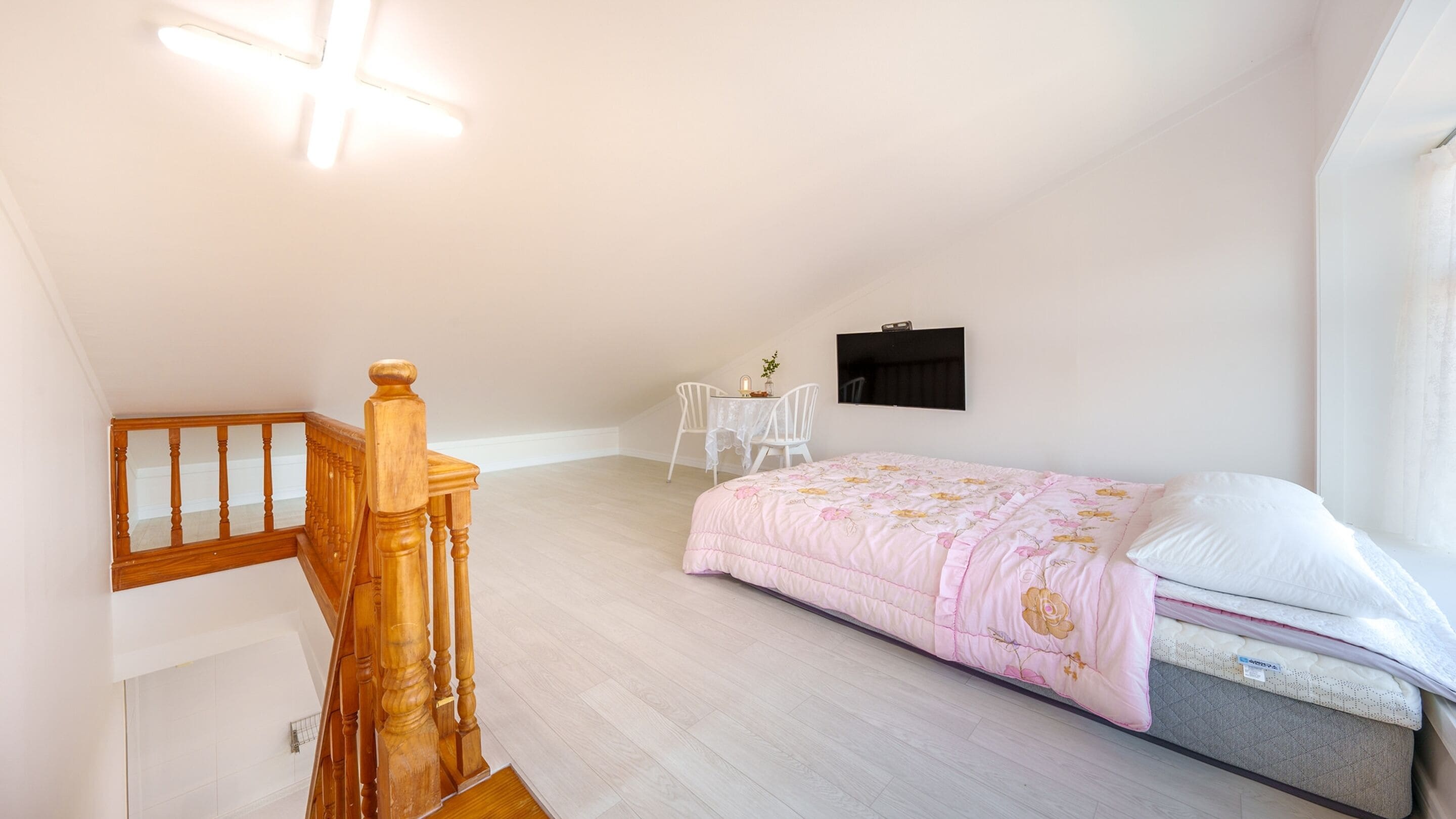 A bedroom with a wooden staircase, a bed with a floral duvet, a small table with chairs, and a TV on the wall.