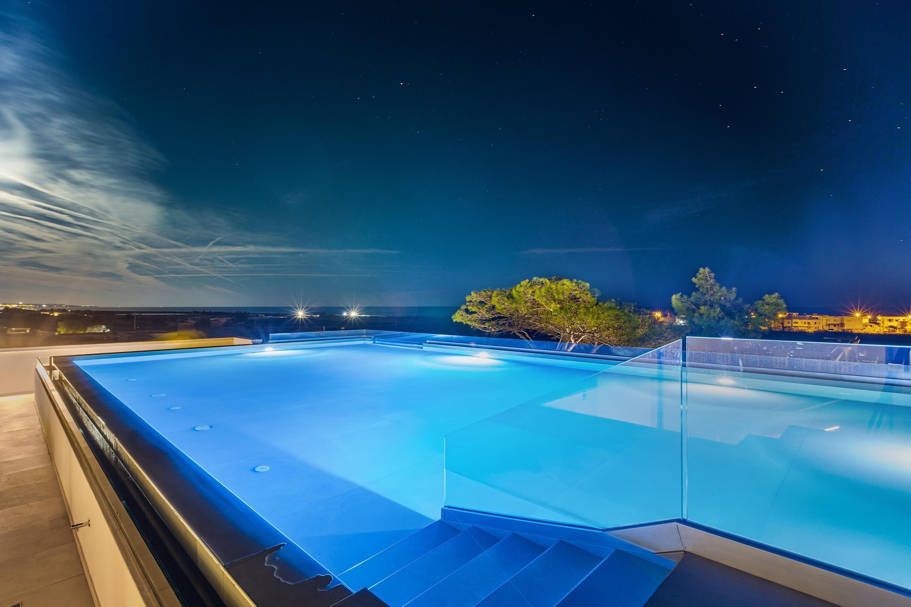 Piscina retangular com água azul cristalina, rodeada por grades de vidro, com céu noturno e estrelas visíveis.