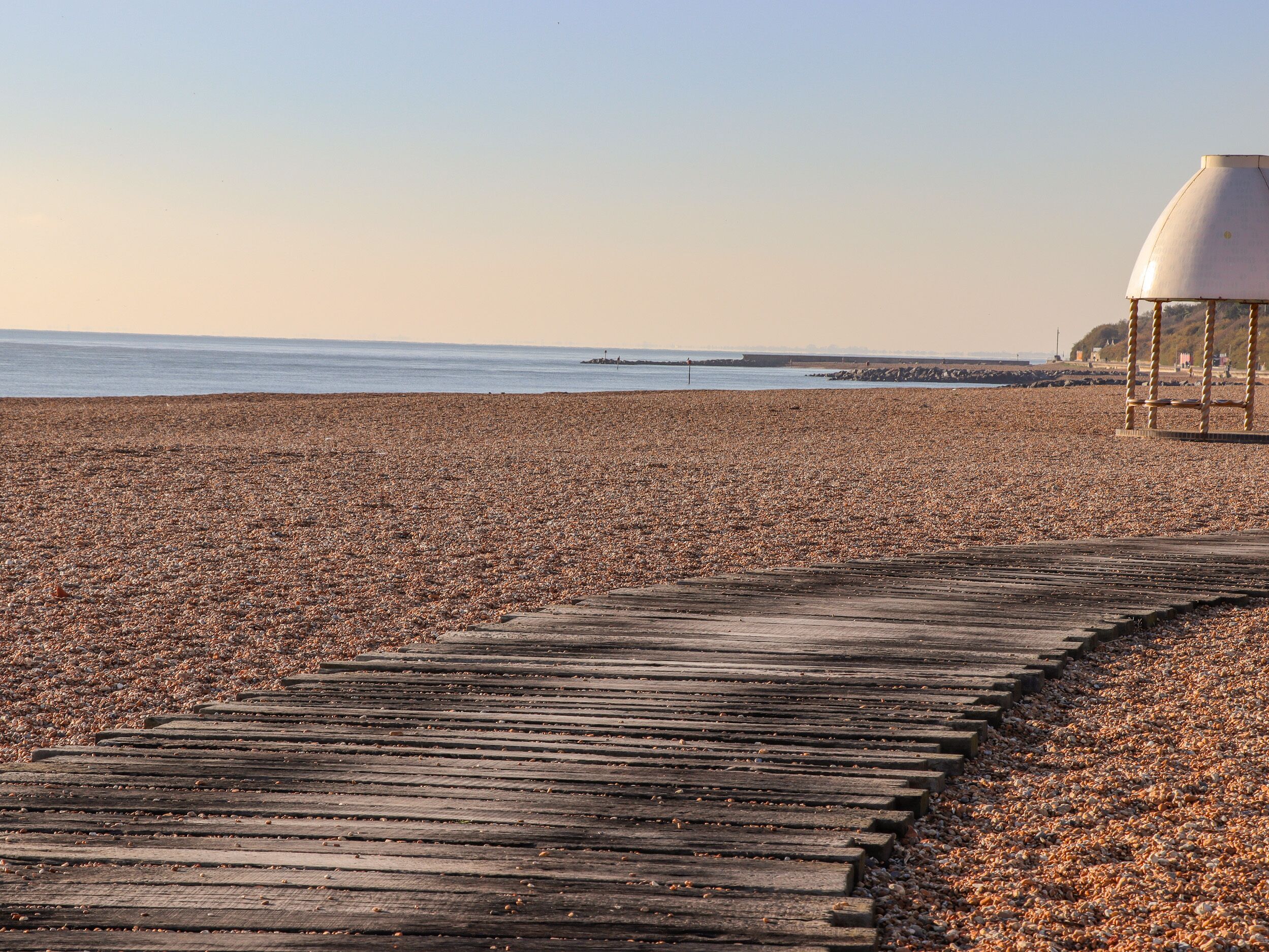 Eine Holzpromenade, die zu einem Kiesstrand und einem Leuchtturm in der Ferne führt.