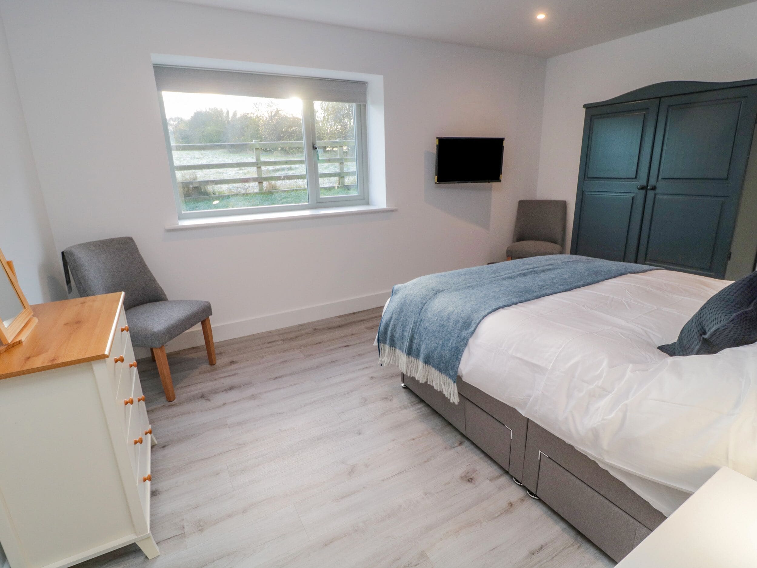 Ein Schlafzimmer mit Bett, Stuhl, Schminktisch, Fernseher und einem Fenster mit Blick auf ein Feld.