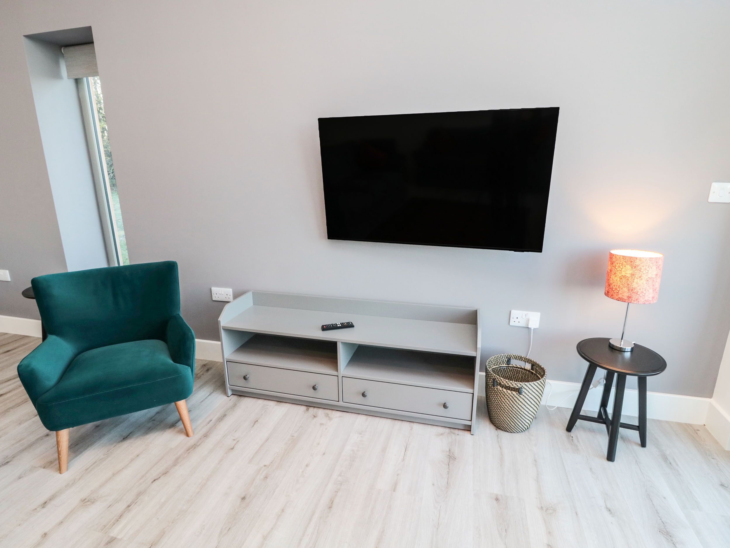 A living room with a teal armchair, a grey entertainment unit, a mounted TV, a lamp, and a small side table.