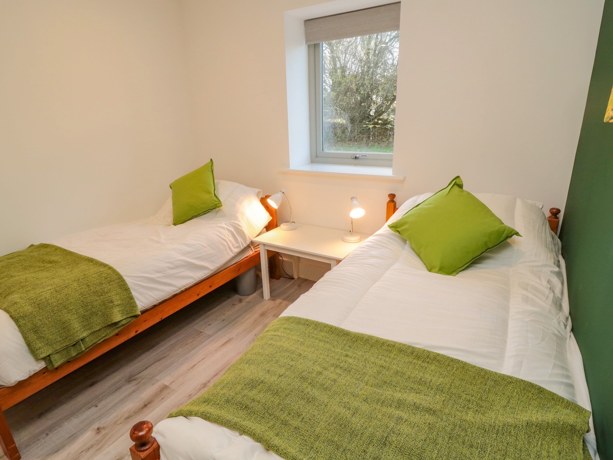 A twin room with two beds, each with a green pillow and a white blanket. There is a small table with a lamp between the beds.