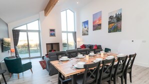Ein moderner Essbereich mit einem Holztisch, gedeckt für sechs Personen, umgeben von schwarzen Stühlen. Im Hintergrund befindet sich eine Couch mit roten Kissen und ein Kamin mit Schornstein. Große Fenster sorgen für natürliches Licht und einen Blick ins Freie.